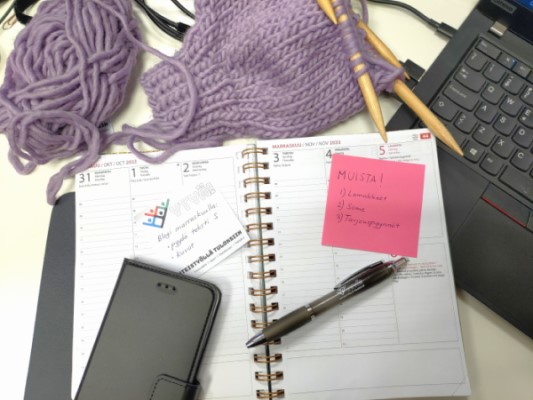 Työpöydällä läppäri, avonainen kalenteri muistilappuineen, puhelin ja neuletyö.
