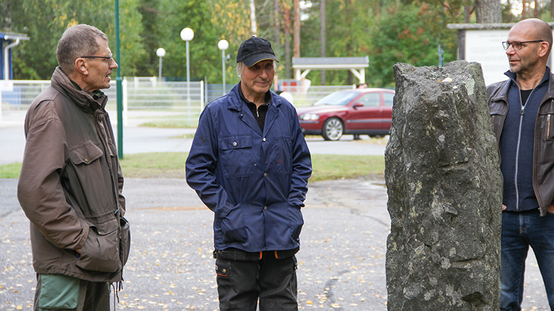 Kolme miestä seisoo kivisen muistomerkin ympärillä.