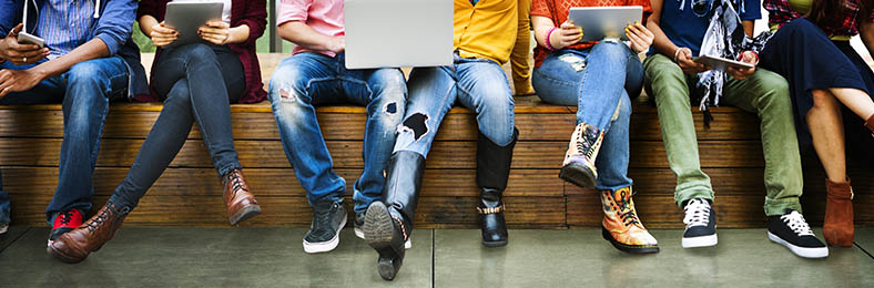 Nuoria ihmisiä istumassa penkillä, käsissä on muun muassa kannettava tietokone, tabletti ja puhelimia.