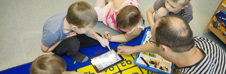 Esiopetuksen lapsia tutkimassa tablettitietokonetta luokkahuoneen lattialla