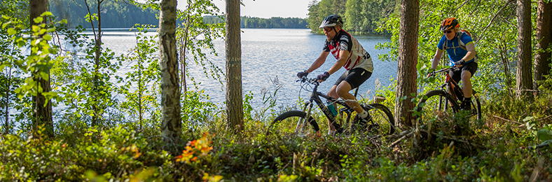 Kaksi maastopyöräilijää kesäisenä päivänä järven rantaa kiertävällä polulla