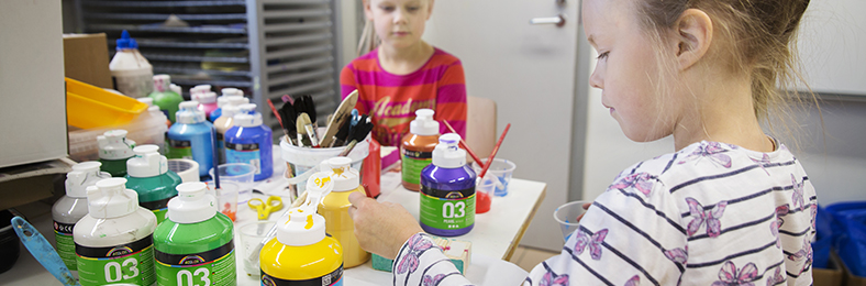 Lapset maalaamassa, pöydällä maalipurkkeja ja siveltimiä