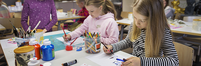 Lapsia luokkahuoneessa piirtämässä ja maalaamassa pulpettien äärellä