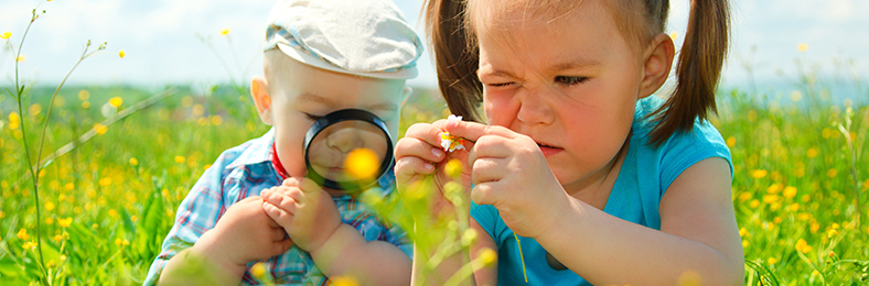 Kaksi lasta aurinkoisella kukkakedolla tutkimassa kasveja suurennuslasilla