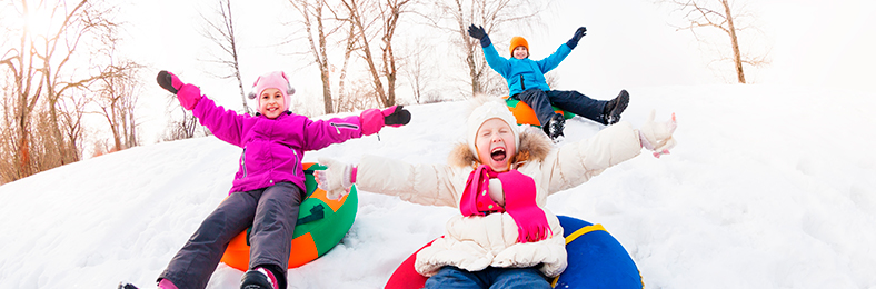Kolme lasta laskemassa talvisena päivänä mäkeä värikkäissä vaatteissa