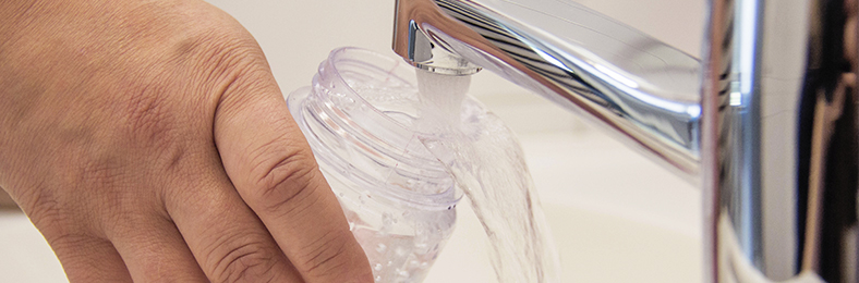 Käsi pitelemässä vesipulloa, hanasta lasketaan vettä pulloon