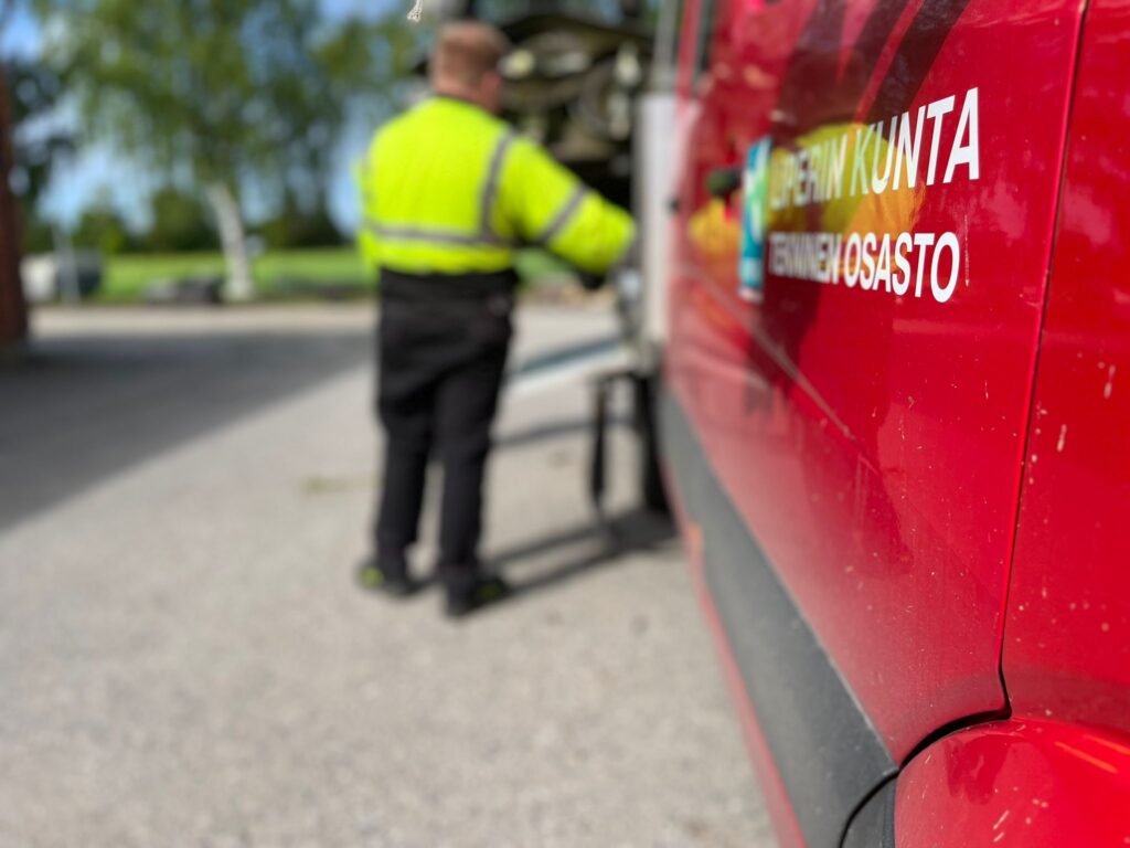 Etualalla työllisyyspalveluiden kuljetusauton ovi, taustalla henkilö huomiovaatteissa.