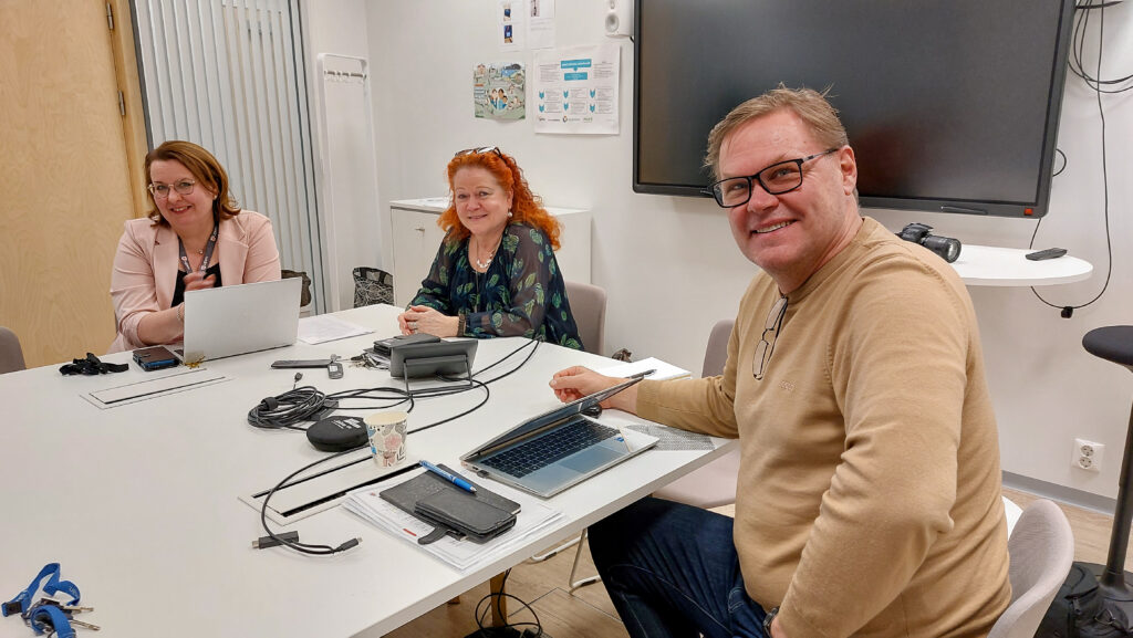 Hyvinvointijohtaja, apulaisrehtori ja rehtori istuvat hymyillen pöydän äärellä kokoustilassa.