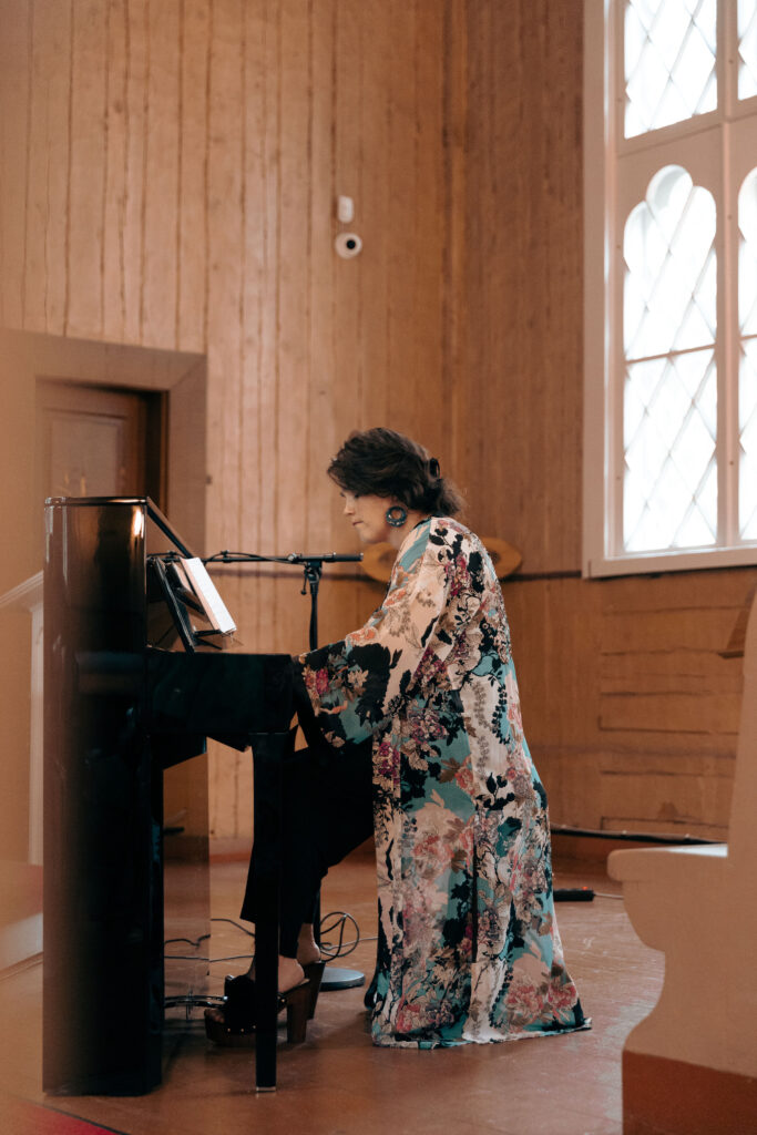 Katja Vanttaja soittaa pianoa. Hänellä on ruskea olkapäämittainen tukka, isot korvakorut, vaalean, turkoosin ja mustan värinen kuviollinen pitkähelmainen vaate, mustat housut ja korolliset kengät.